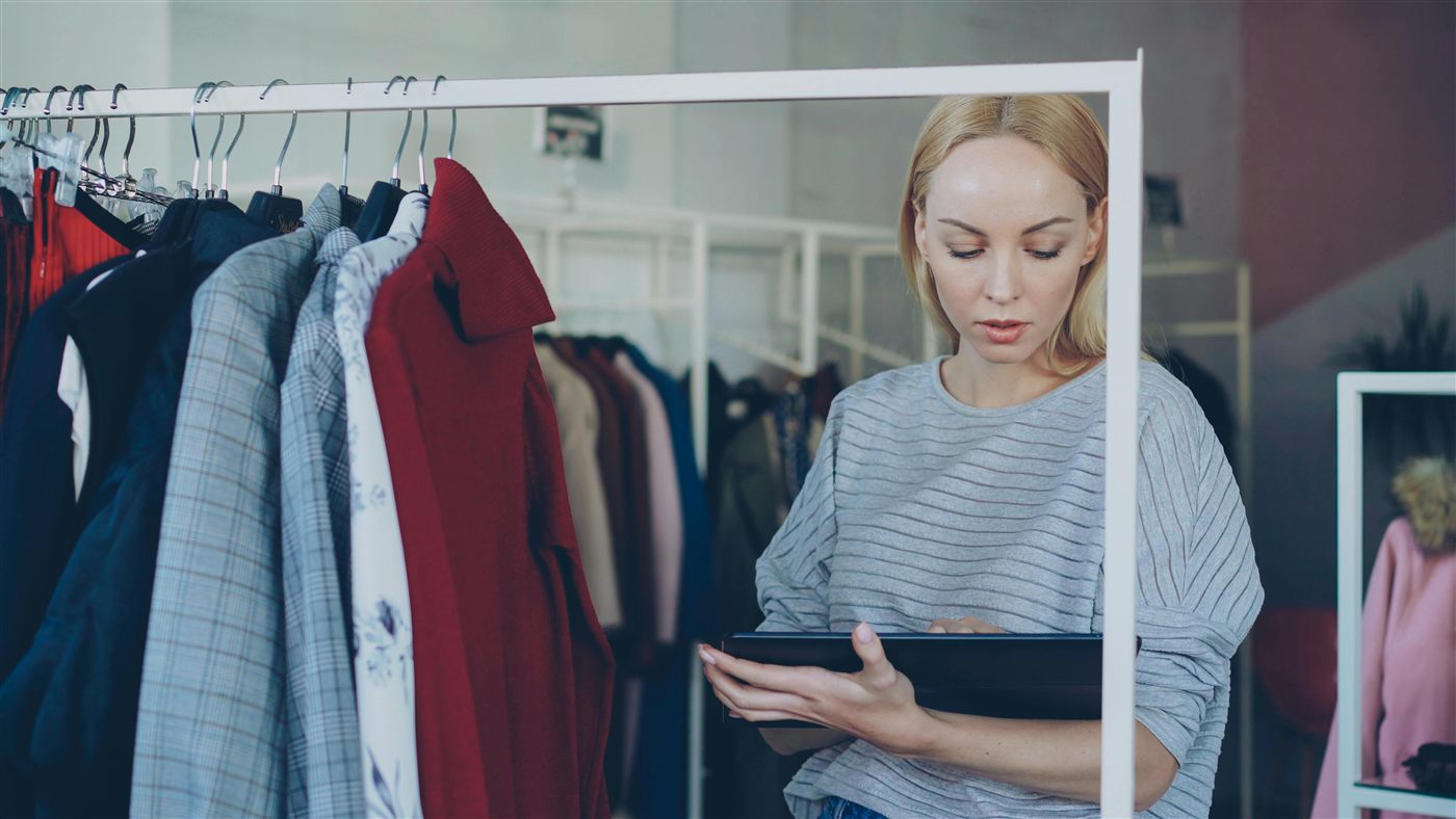 Retail assistant checking clothing stock on a tablet beside a product rail.